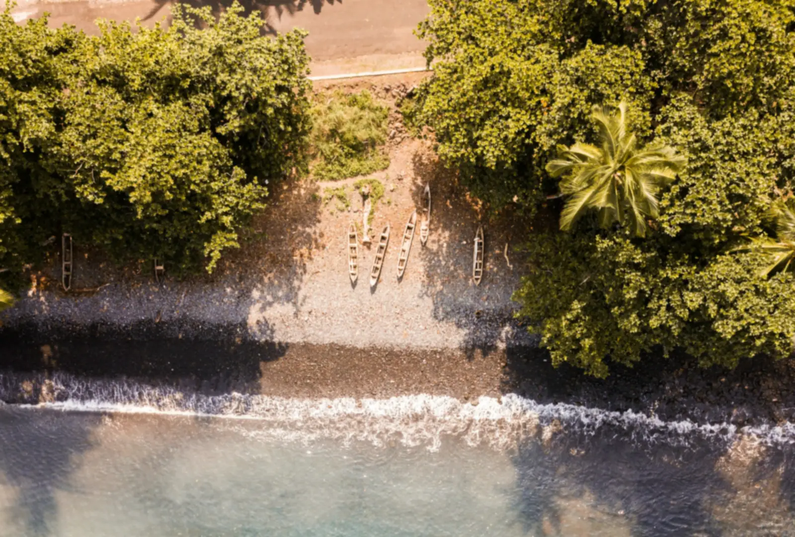Ein Strand mit dunklem Sand auf São Tomé von oben, mit Palmen, dichtem Grün und klarem türkisfarbenem Wasser